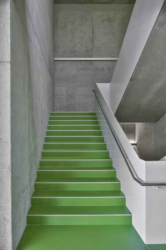 Das Bild zeigt ein Treppenhaus der HfK Ulm mit Sichtbetonwänden und grüner Treppe